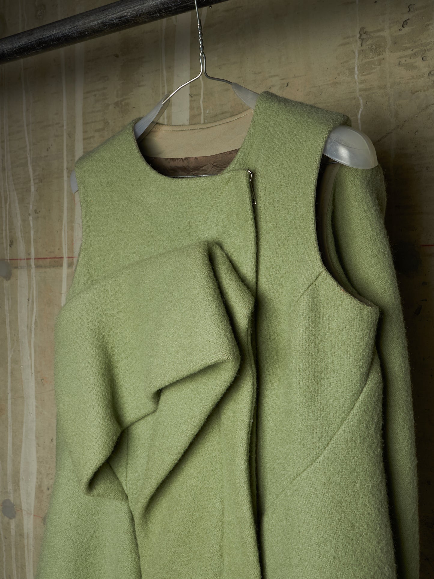 A sage green wool-blend vest with an asymmetrical draped front detail and a central zipper, hanging on a wire hanger against a textured concrete wall.