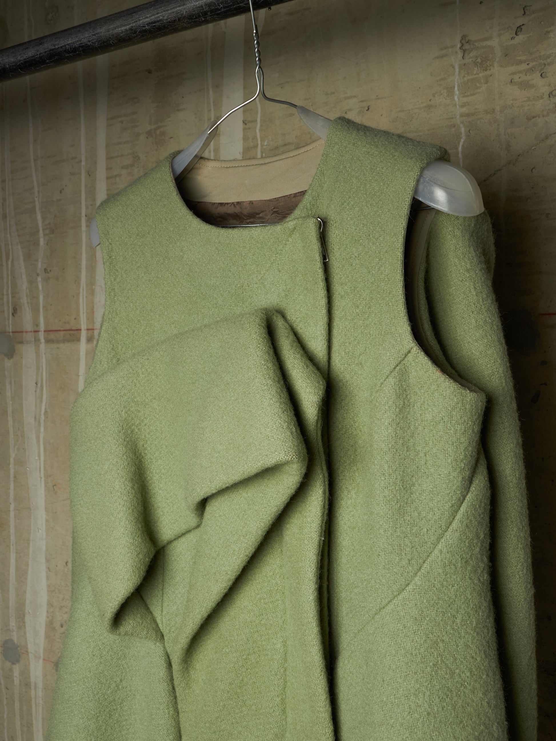 A sage green wool-blend vest with an asymmetrical draped front detail and a central zipper, hanging on a wire hanger against a textured concrete wall.