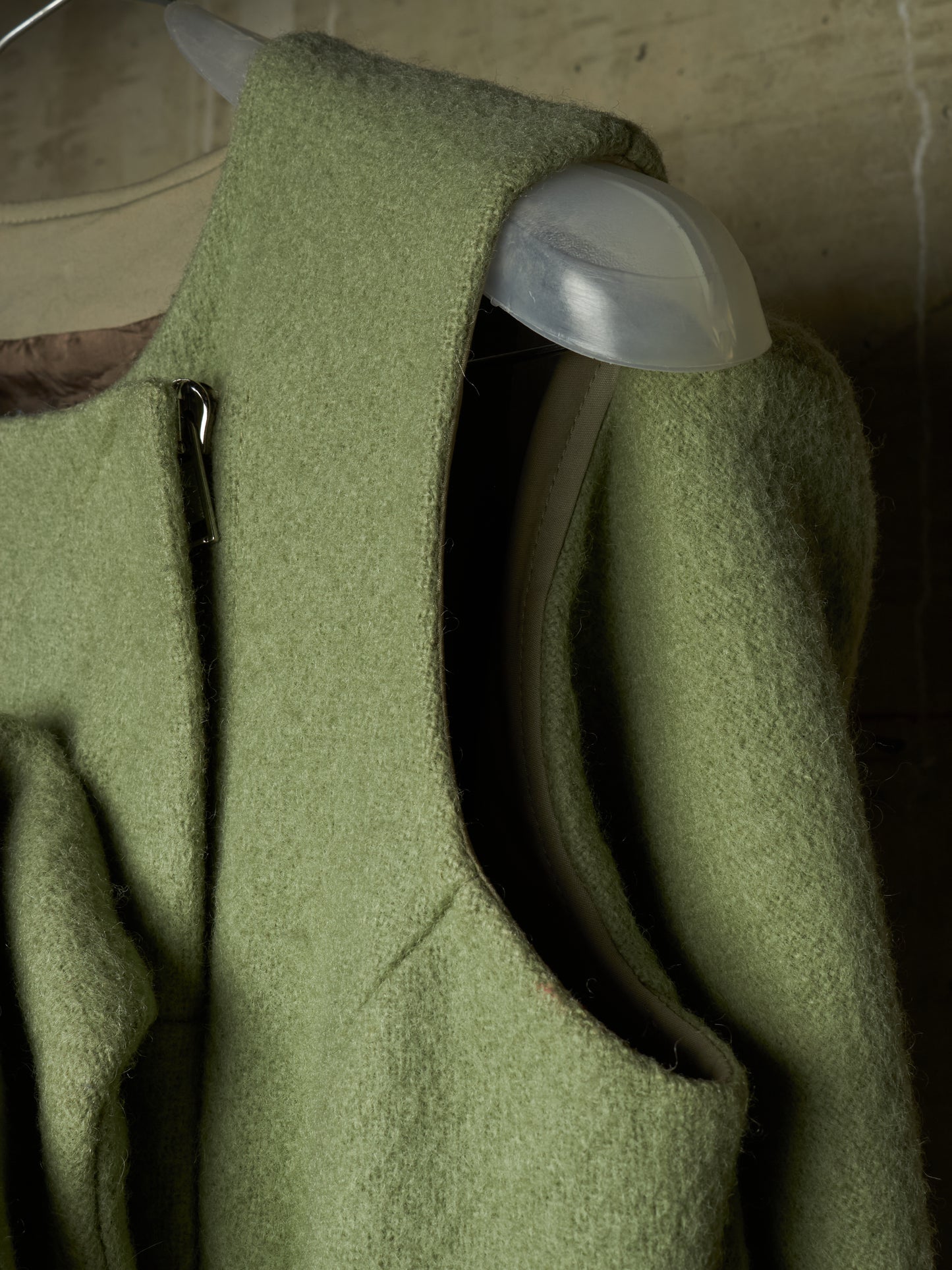 Close-up of a sage green wool coat hanging on a clear plastic hanger against a rustic wooden background.