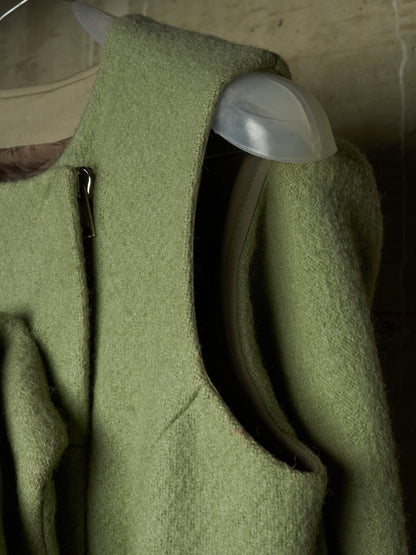Close-up of a sage green wool coat hanging on a clear plastic hanger against a rustic wooden background.