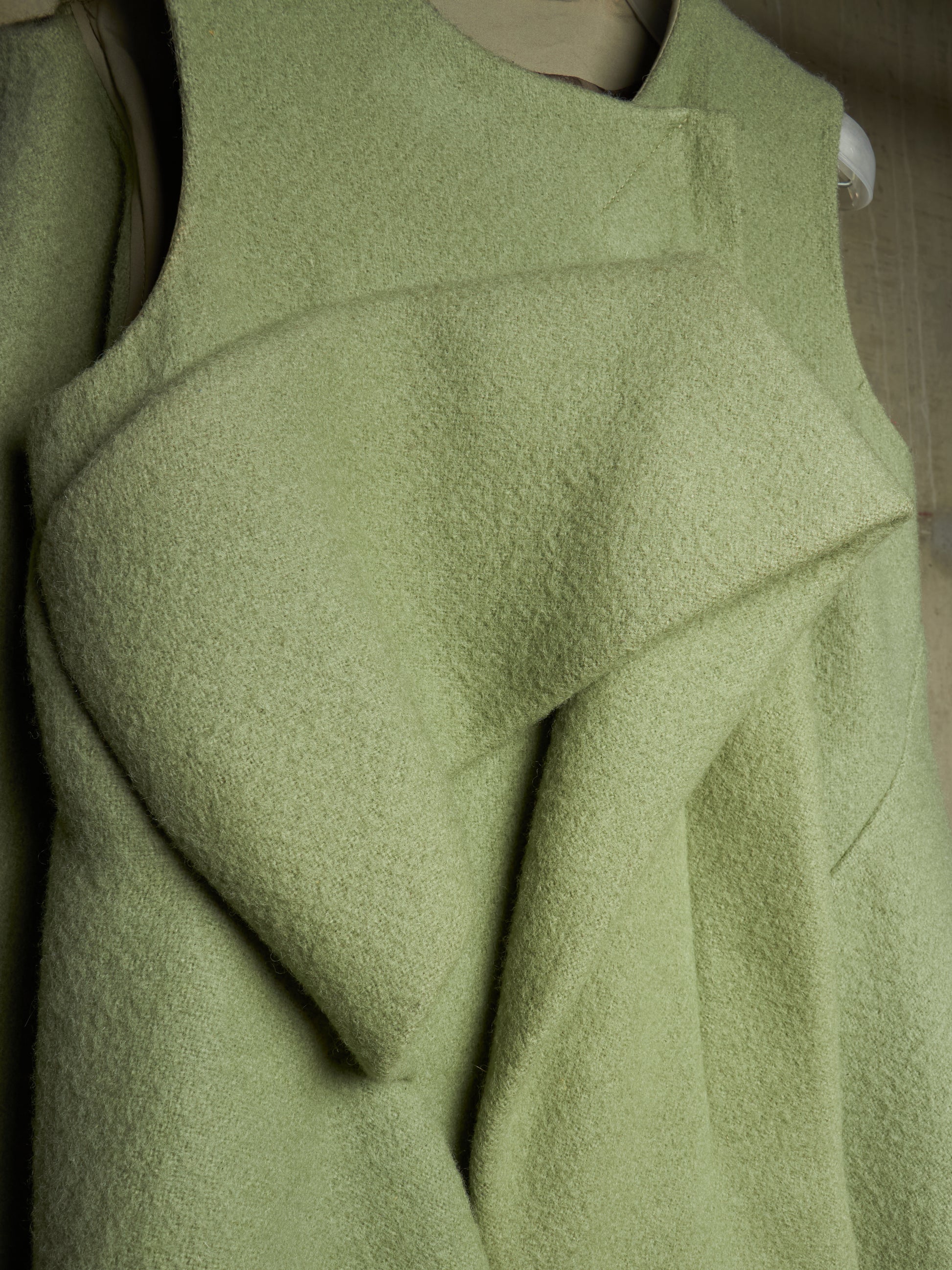 A close-up of a light sage green textured garment, likely a wool sweater or coat, featuring draped, asymmetrical folds and a sleeveless design.