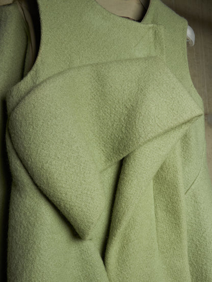 A close-up of a light sage green textured garment, likely a wool sweater or coat, featuring draped, asymmetrical folds and a sleeveless design.