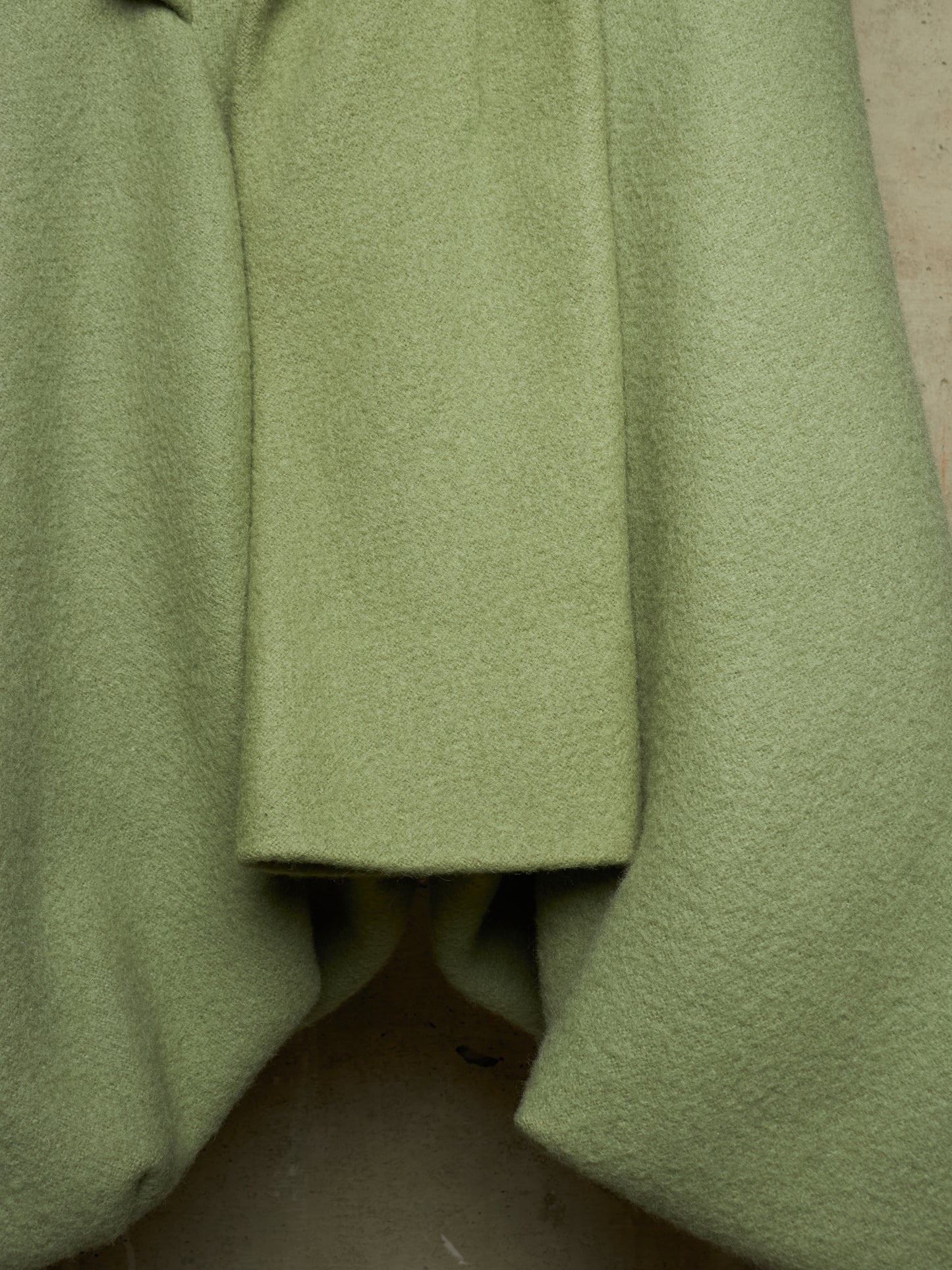 A close-up of a light olive green wool coat with a textured bouclé fabric, showing the draped folds and lapel detail.