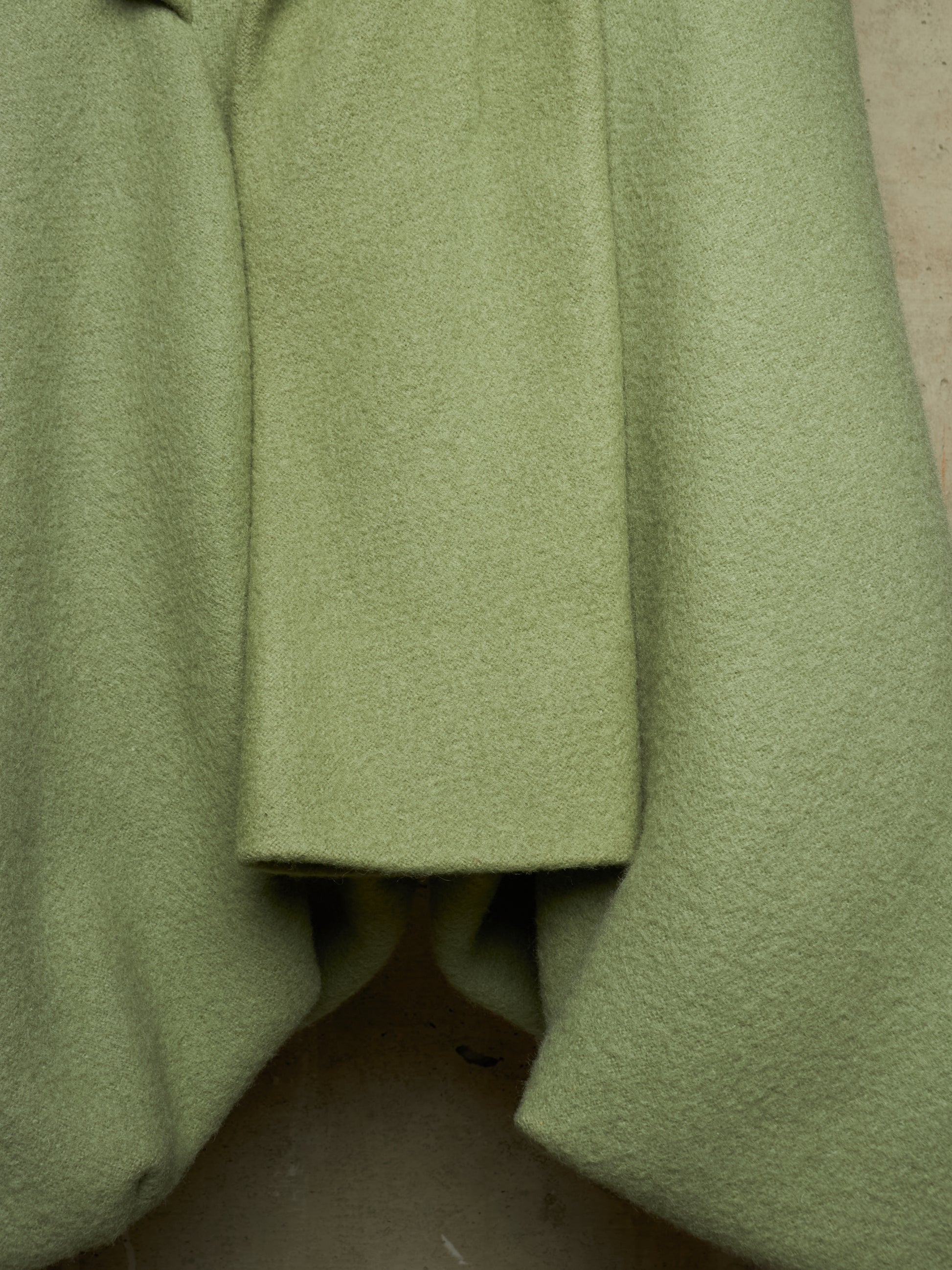 A close-up of a light olive green wool coat with a textured bouclé fabric, showing the draped folds and lapel detail.