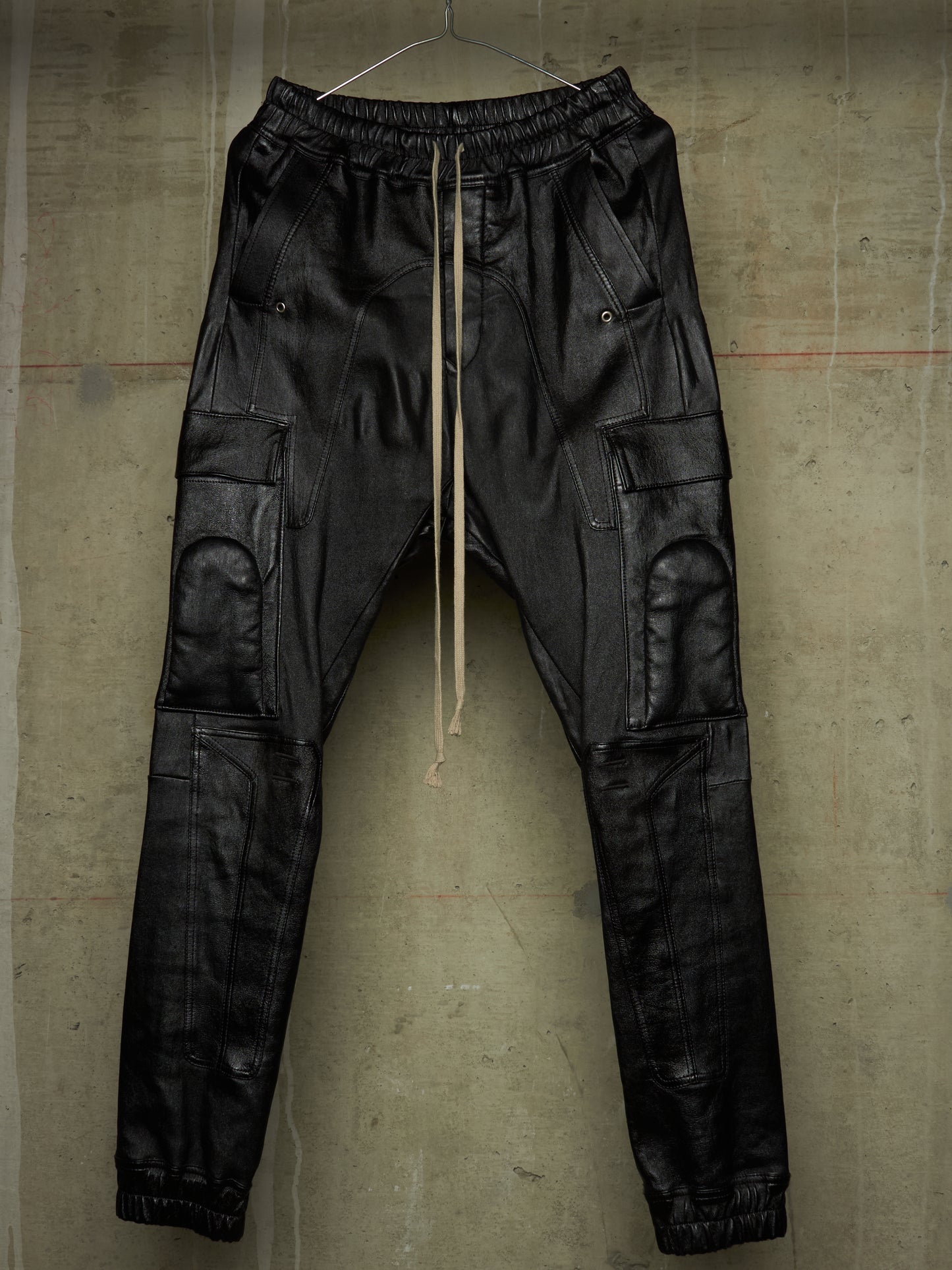 Black faux leather cargo pants with an elastic waistband, beige drawstring, and multiple utility pockets, hanging against a textured concrete wall.