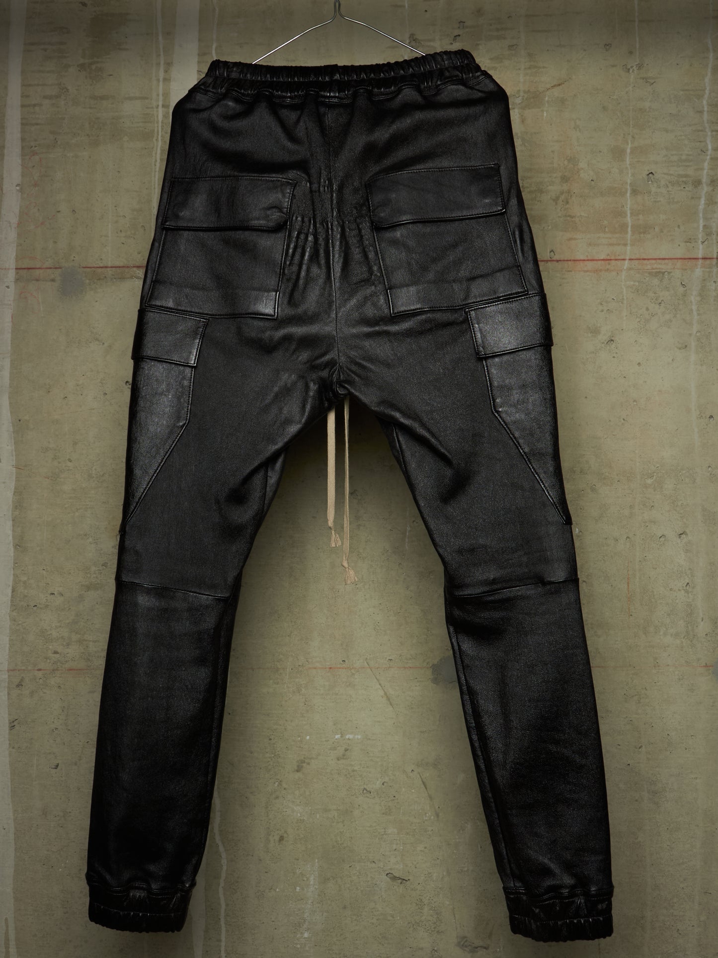 Black leather cargo pants with an elastic waistband, hanging against a textured concrete wall.
