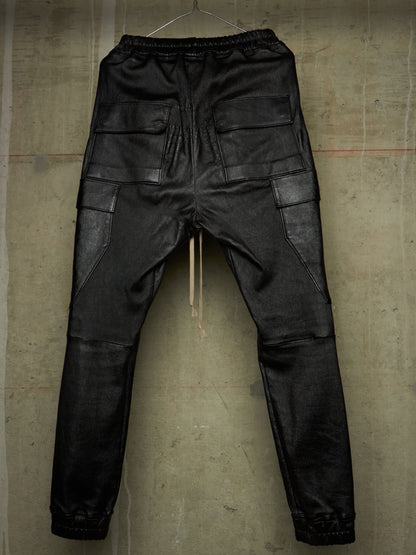 Black leather cargo pants with an elastic waistband, hanging against a textured concrete wall.