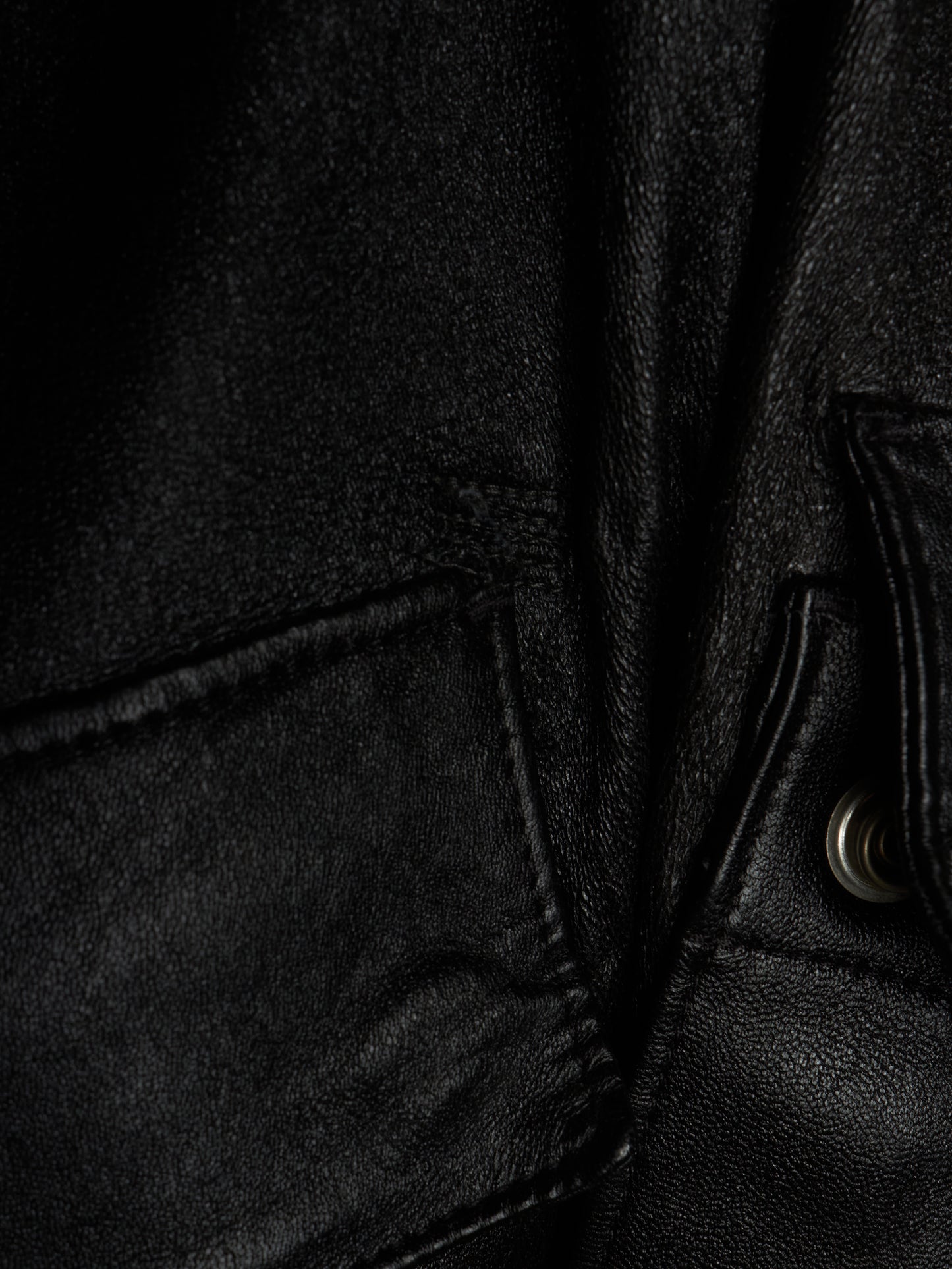 Close-up detail of a black leather jacket showing the texture of the leather, a pocket seam, and a silver metal snap button.