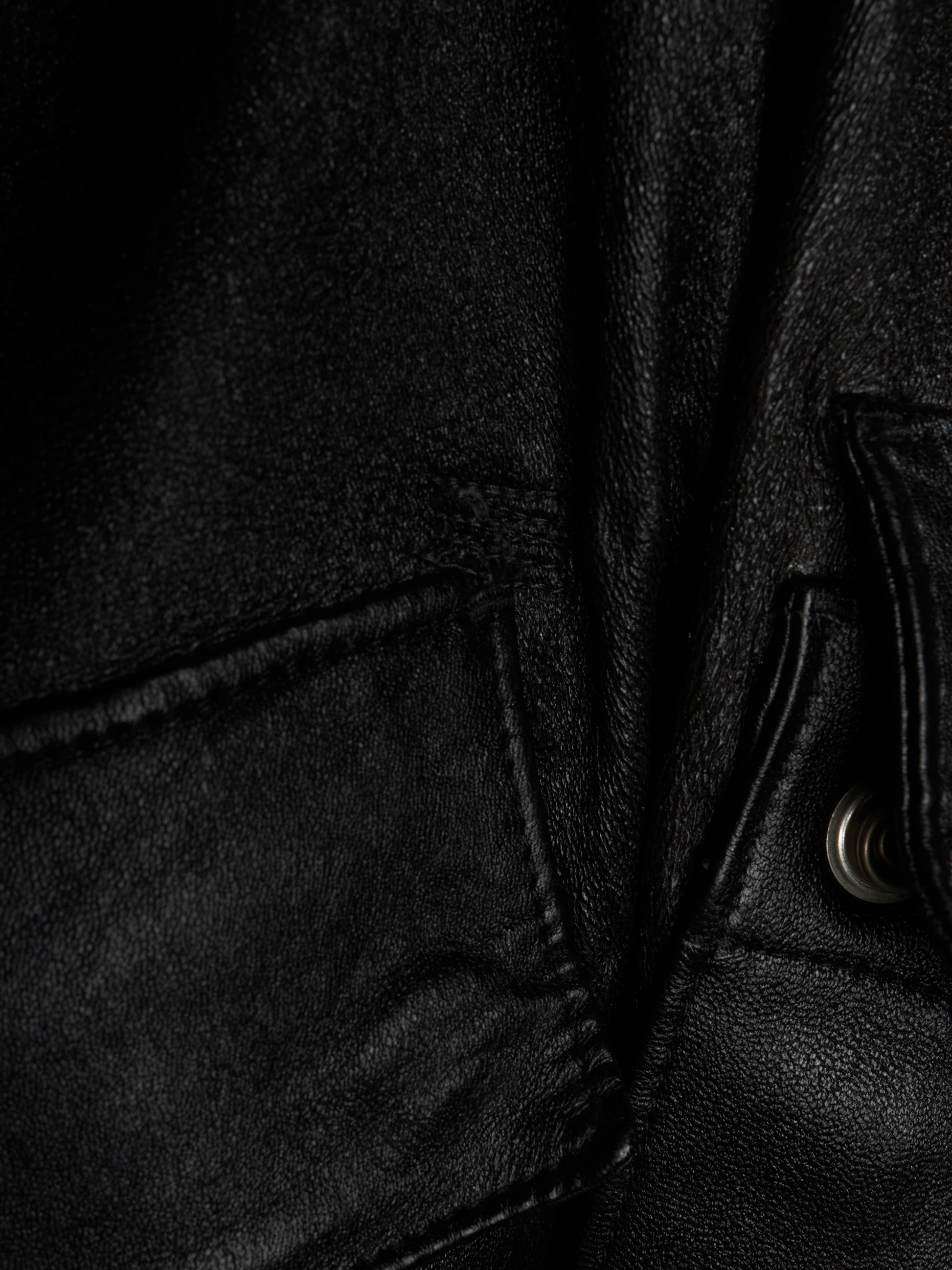 Close-up detail of a black leather jacket showing the texture of the leather, a pocket seam, and a silver metal snap button.