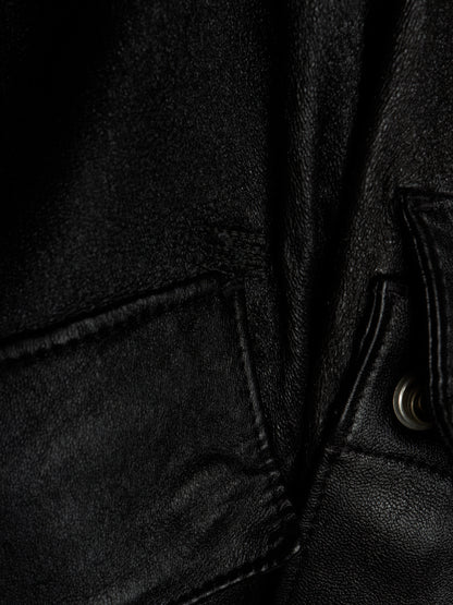 Close-up detail of a black leather jacket showing the texture of the leather, a pocket seam, and a silver metal snap button.