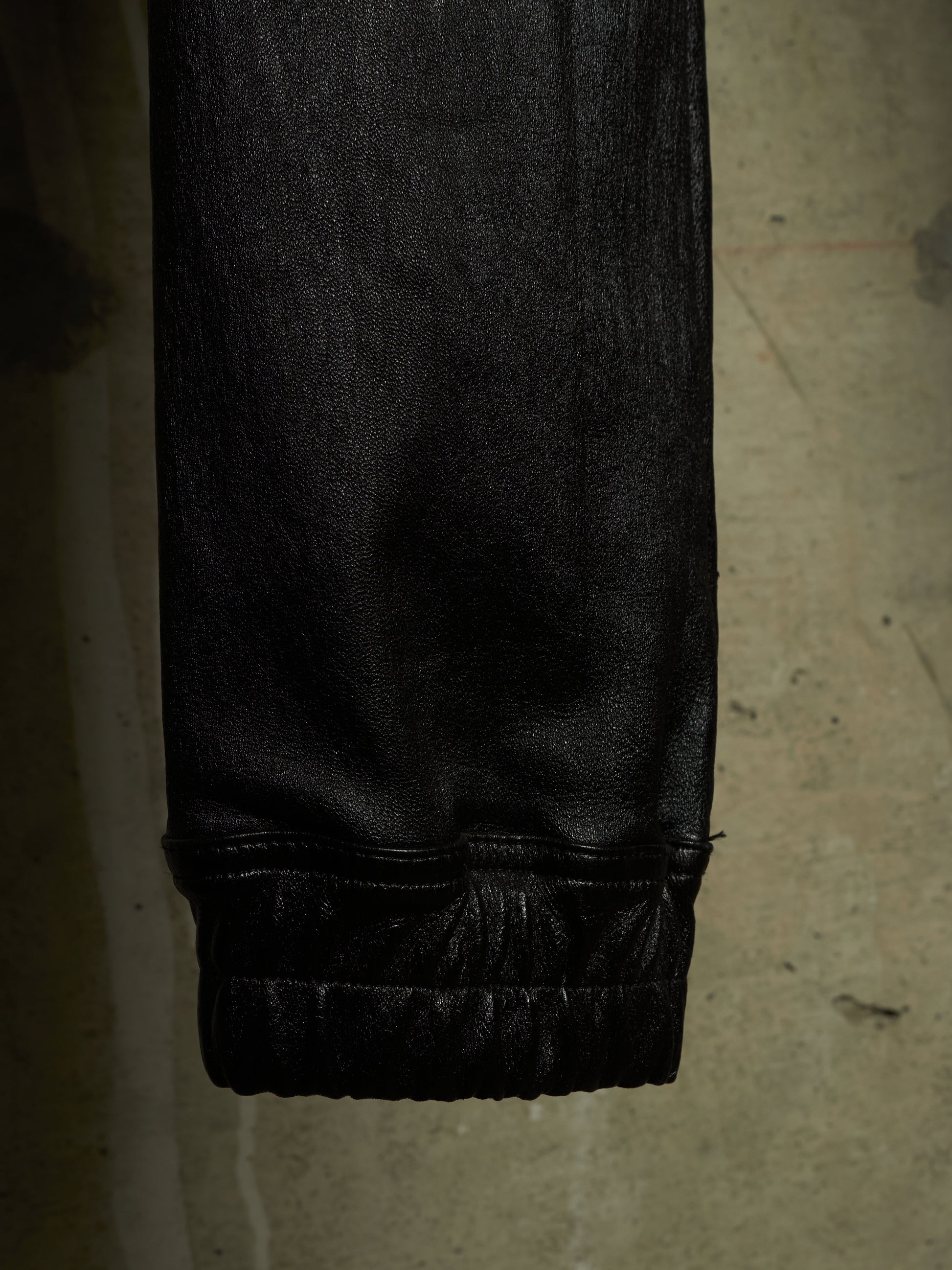 Close-up of a black textured garment, likely leather or faux leather pants, showing a gathered elastic cuff against a concrete floor background.