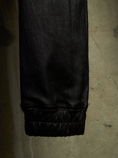 Close-up of a black textured garment, likely leather or faux leather pants, showing a gathered elastic cuff against a concrete floor background.