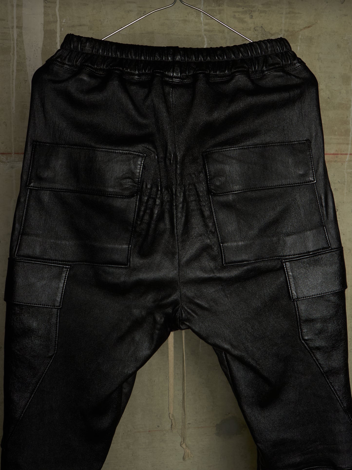 Black faux leather cargo pants with an elastic waistband, shown from the back on a hanger against a textured concrete wall.