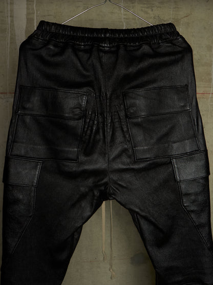 Black faux leather cargo pants with an elastic waistband, shown from the back on a hanger against a textured concrete wall.