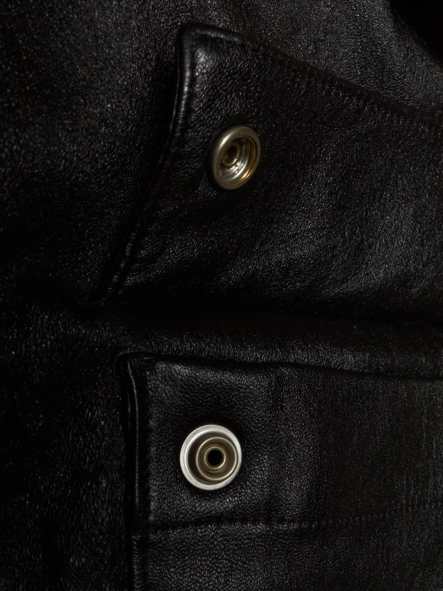 Close-up of a black leather jacket showing two silver metal snap buttons on angled flap pockets.