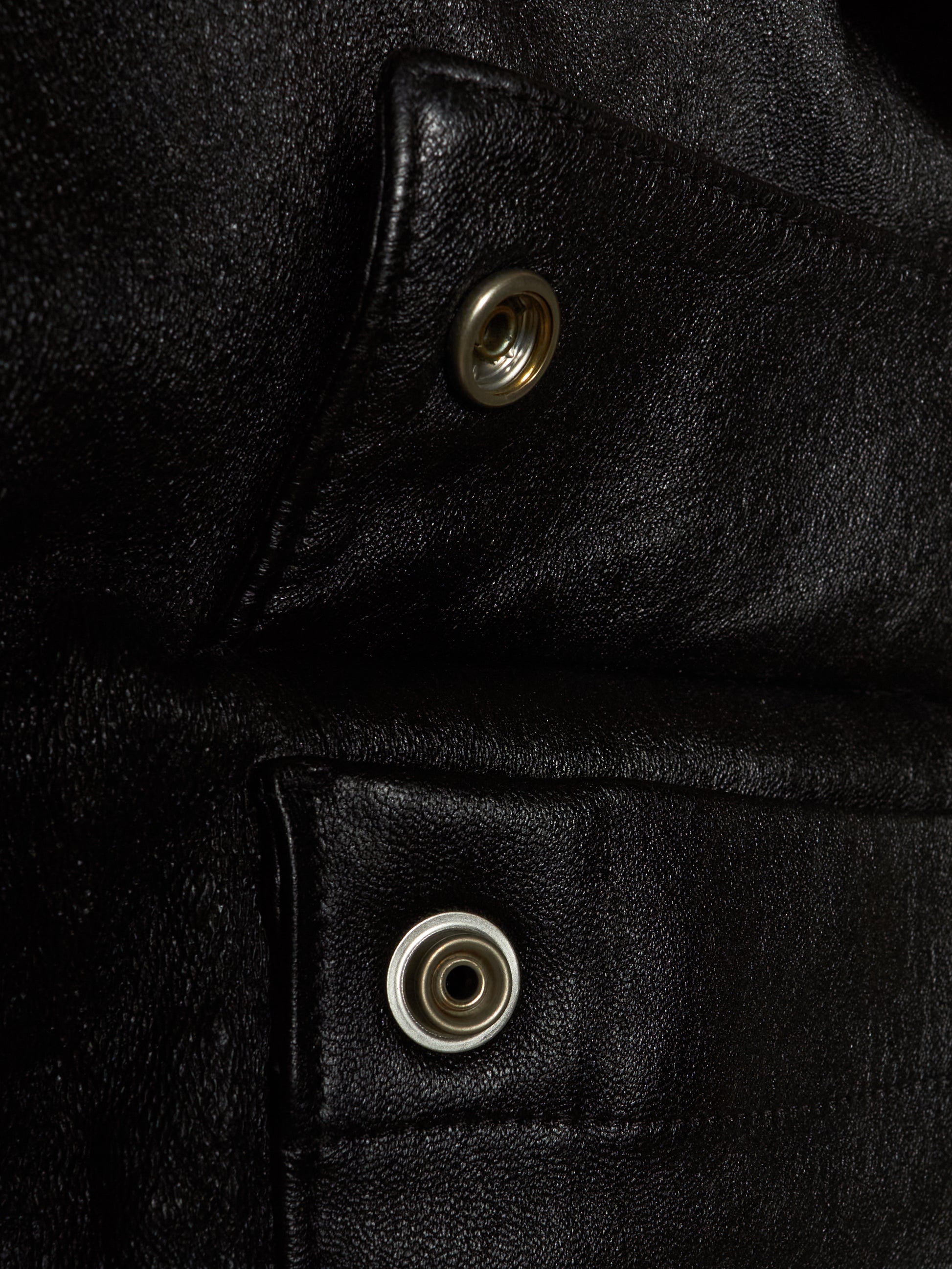 Close-up of a black leather jacket showing two silver metal snap buttons on angled flap pockets.