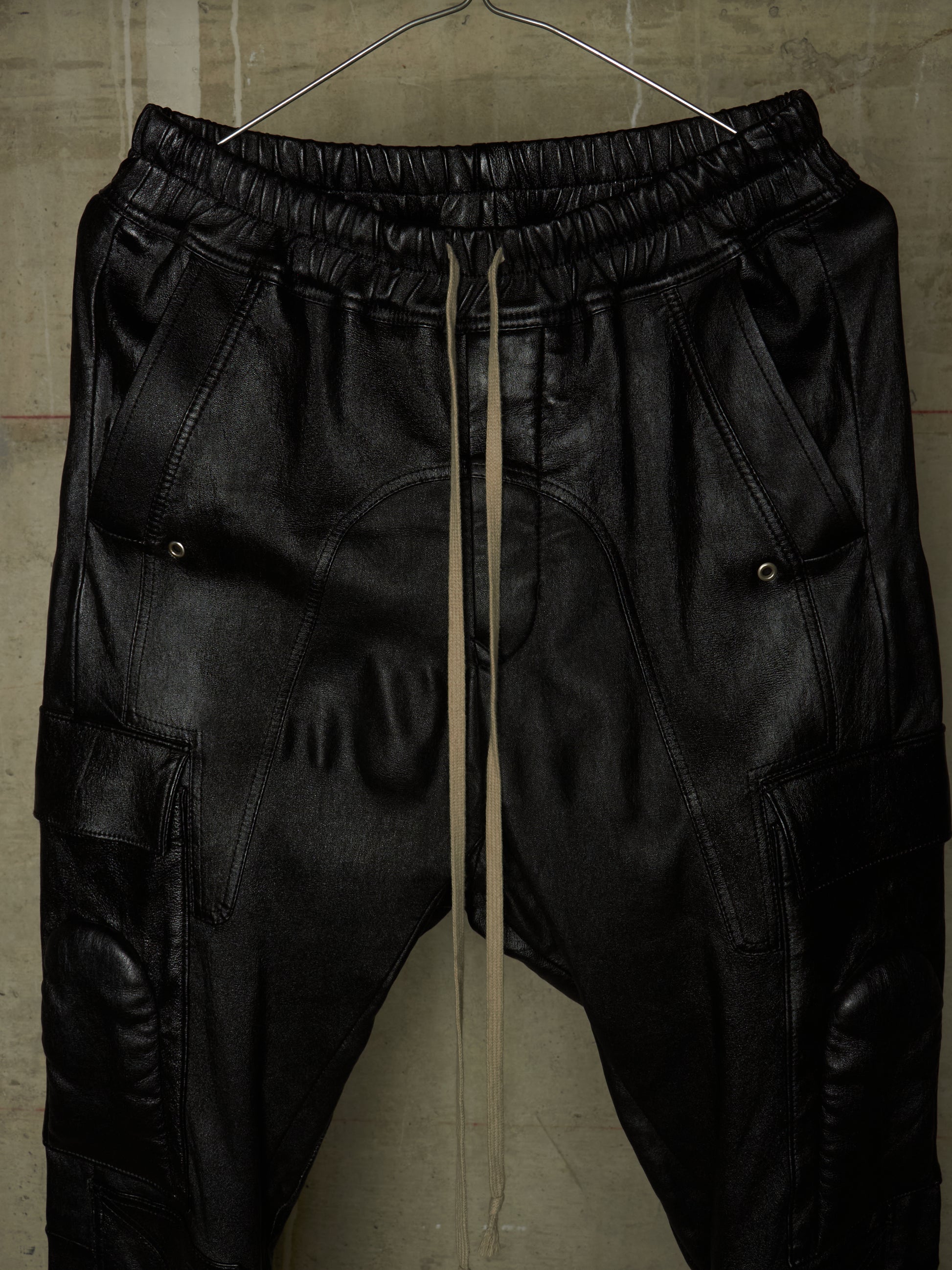 Black leather jogger pants with an elastic waistband, drawstring, and cargo pockets, hanging on a wire hanger against a textured concrete wall.