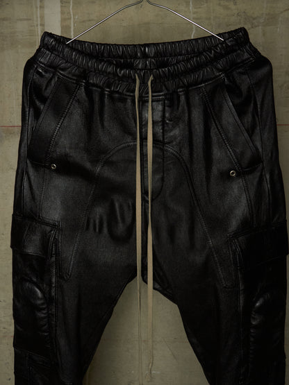 Black leather jogger pants with an elastic waistband, drawstring, and cargo pockets, hanging on a wire hanger against a textured concrete wall.