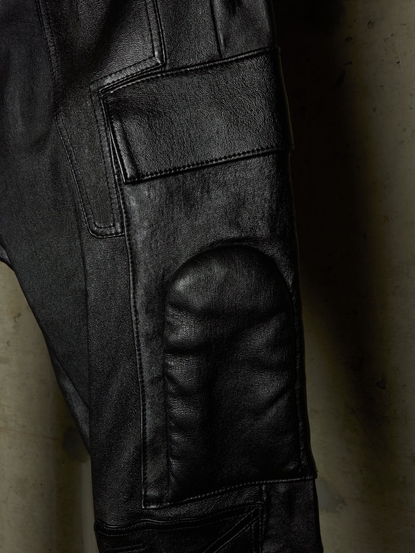 Close-up of black leather pants showing the textured leg, cargo pocket, and reinforced knee patch against a dark background.