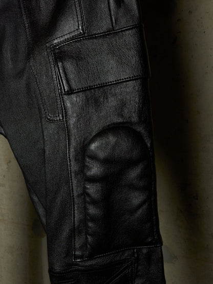 Close-up of black leather pants showing the textured leg, cargo pocket, and reinforced knee patch against a dark background.