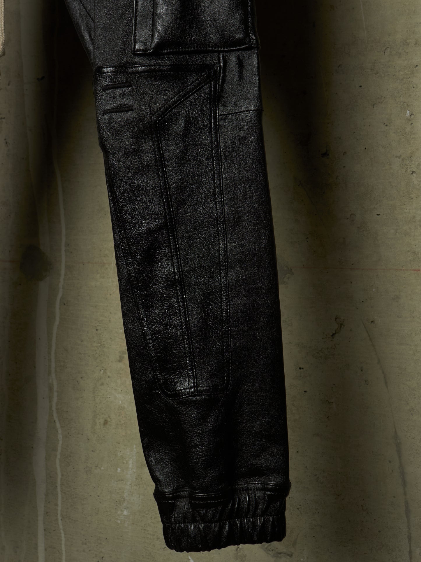 Close-up of a black leather garment, likely a jacket or pants leg, hanging against a textured concrete wall. The fabric shows visible seams, a cuff, and a dark elastic band.