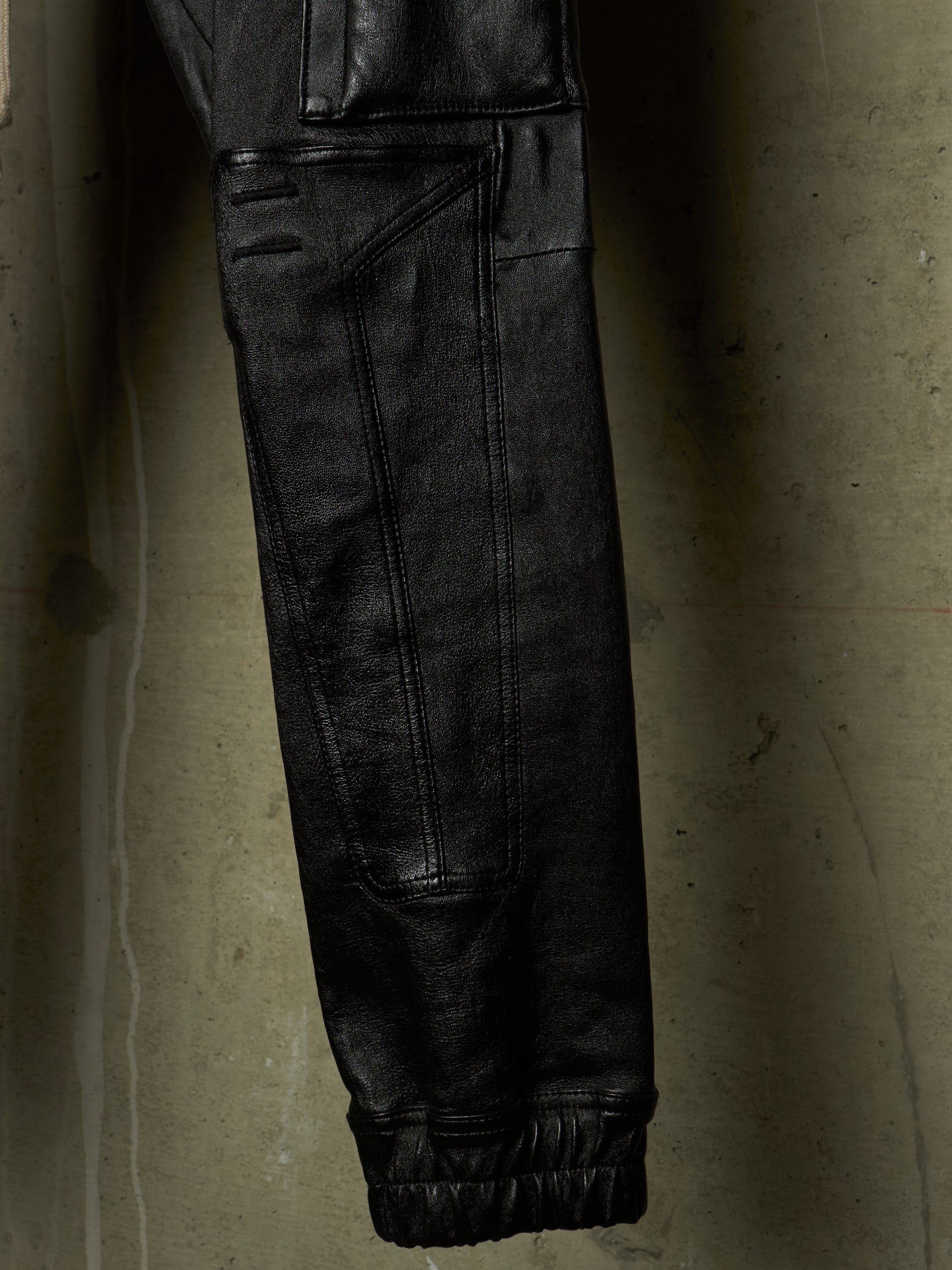 Close-up of a black leather garment, likely a jacket or pants leg, hanging against a textured concrete wall. The fabric shows visible seams, a cuff, and a dark elastic band.