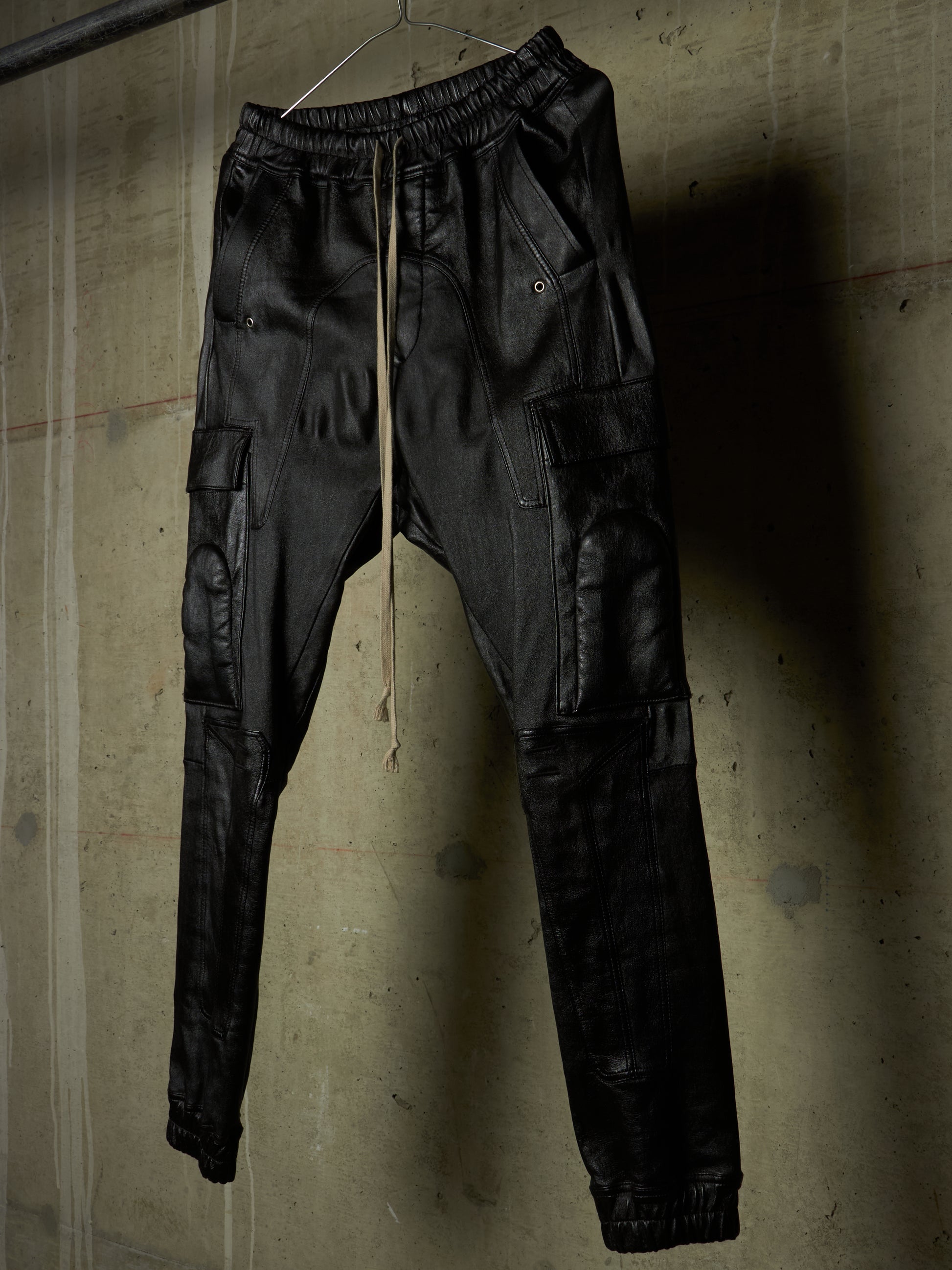 Black leather cargo jogger pants hanging on a wire hanger against a textured concrete wall.