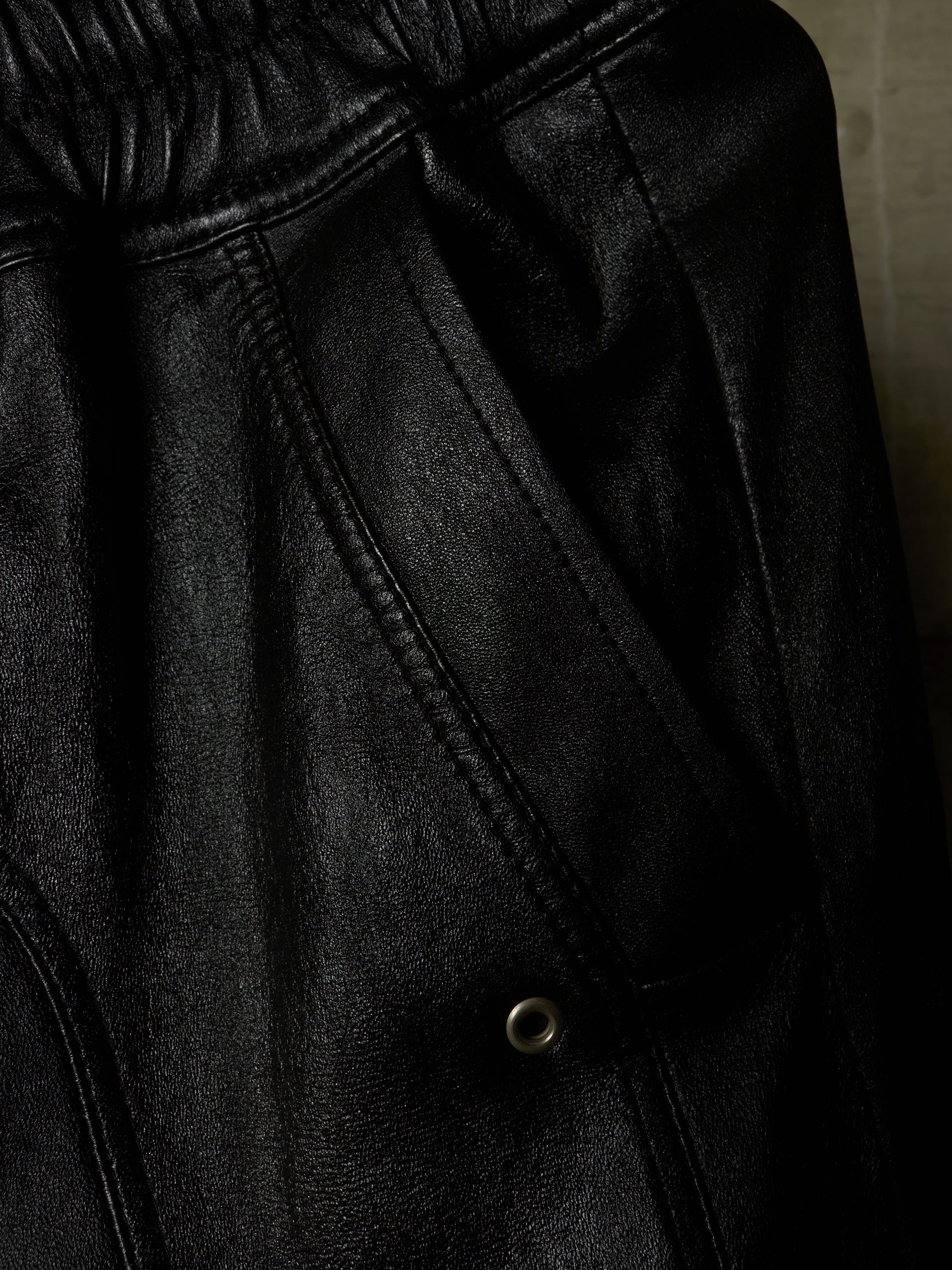 Close-up detail shot of black faux leather pants showing the elastic waistband, stitching, and a metal snap button.