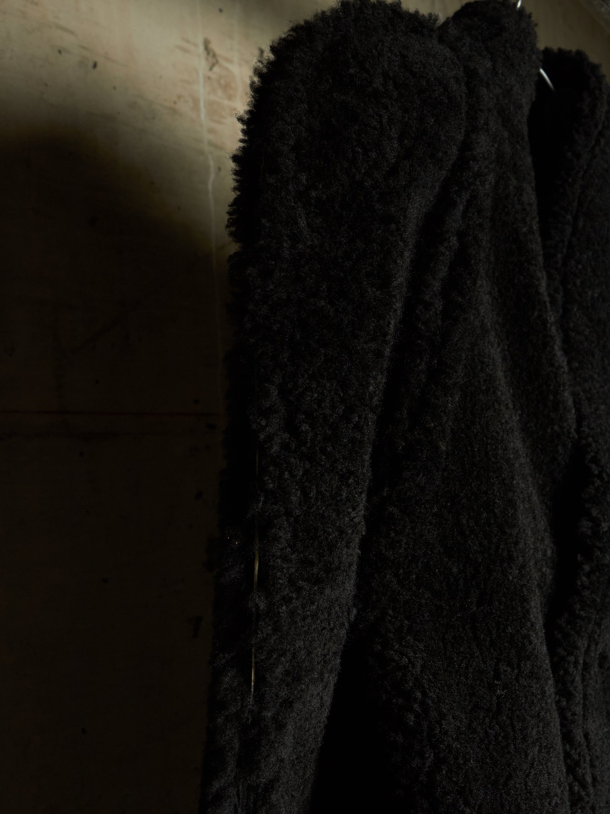 A close-up of a black textured teddy coat or jacket hanging against a dimly lit beige wall.