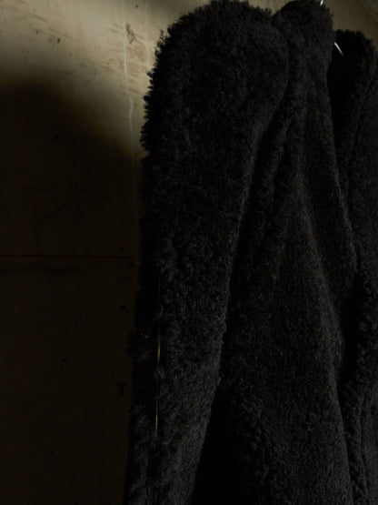 A close-up of a black textured teddy coat or jacket hanging against a dimly lit beige wall.