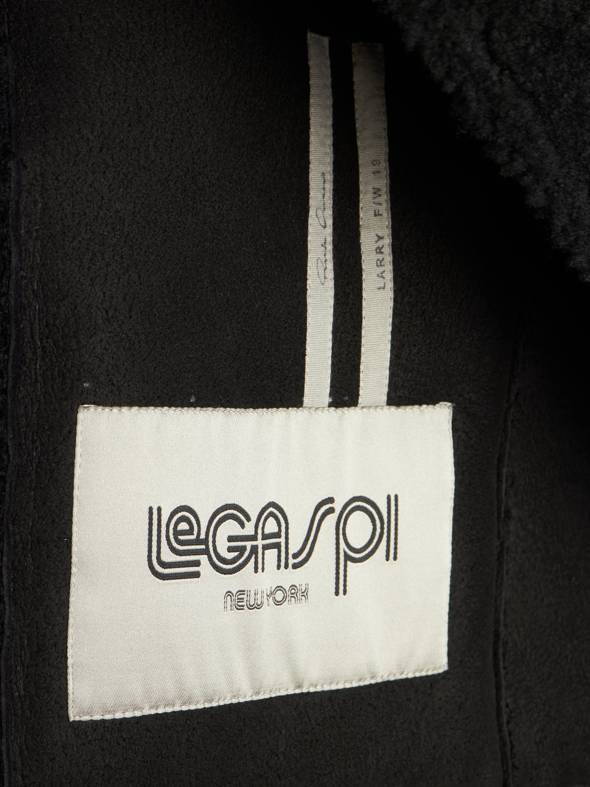 Close-up of a black garment interior showing a white sewn-in label that reads 'LEGASP1 NEW YORK' and a striped inner tag.