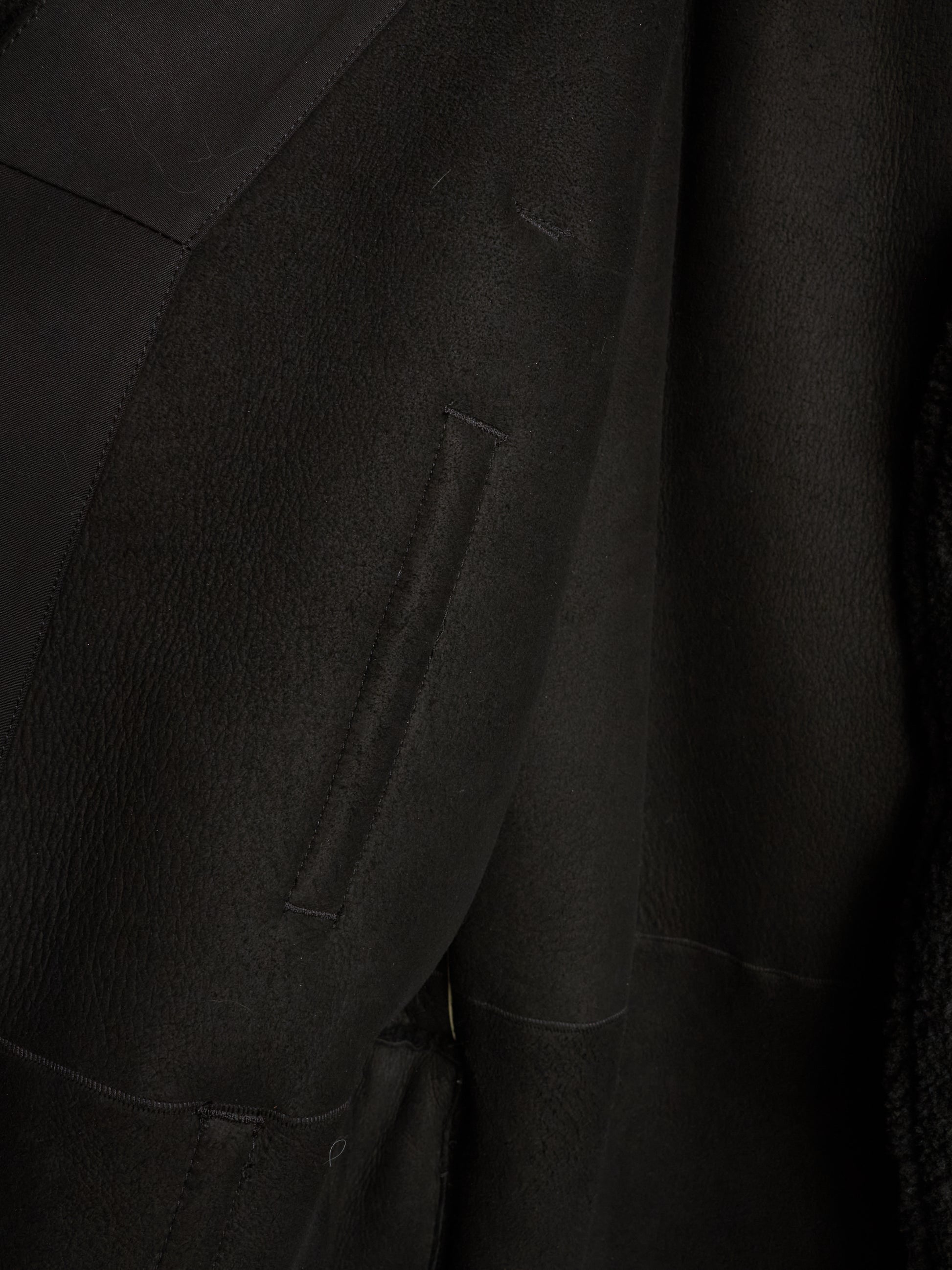 Close-up detail of a black textured jacket or coat showing a vertical welt pocket and fabric texture.