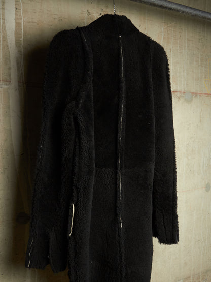 A black long-sleeve coat with mixed textures, including a fuzzy sherpa-like panel and smoother fabric, hanging against a textured beige wall.
