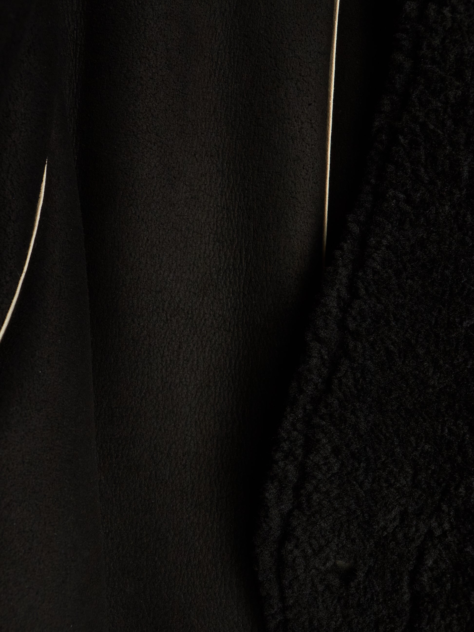 Close-up detail of a black textured garment, showing a smooth matte fabric panel next to a chunky knit or bouclé texture, with thin light-colored piping lines visible.