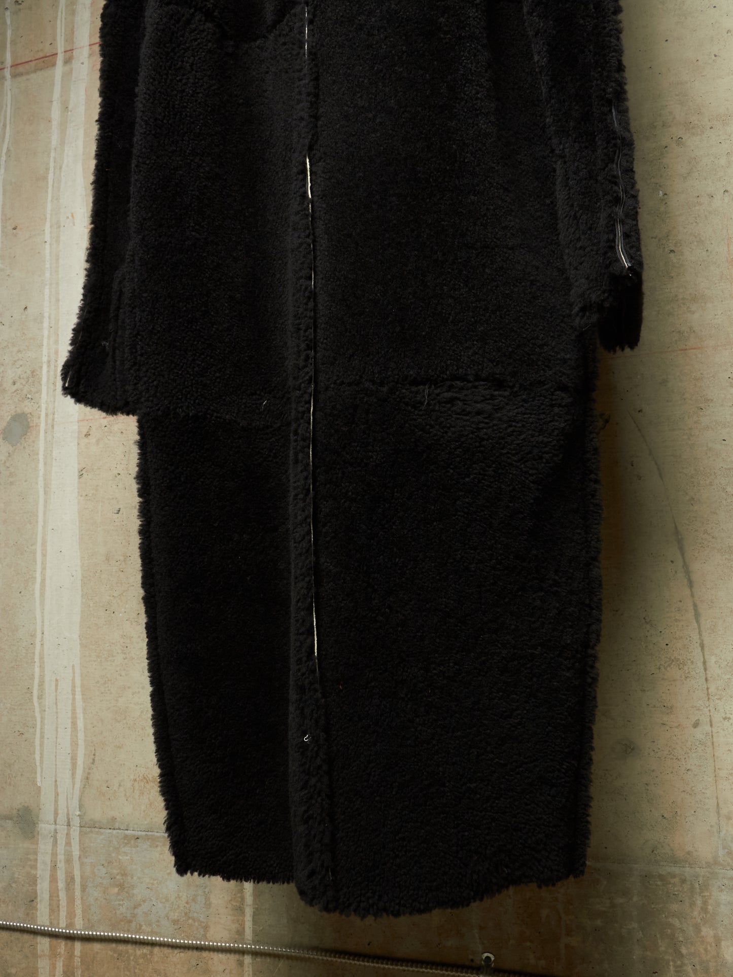 A black fuzzy teddy coat with a front zipper, shown hanging against a textured concrete wall.
