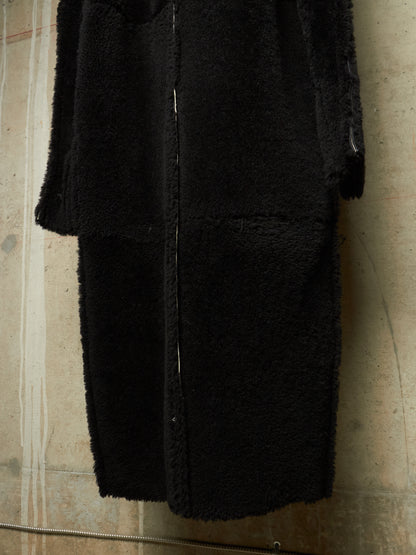 A black fuzzy teddy coat with a front zipper, shown hanging against a textured concrete wall.
