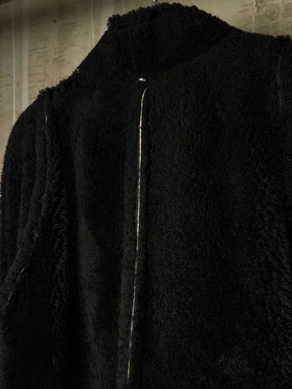 Close-up of a black textured faux-fur or sherpa jacket with a high collar and a vertical silver zipper, hanging against a light wall.