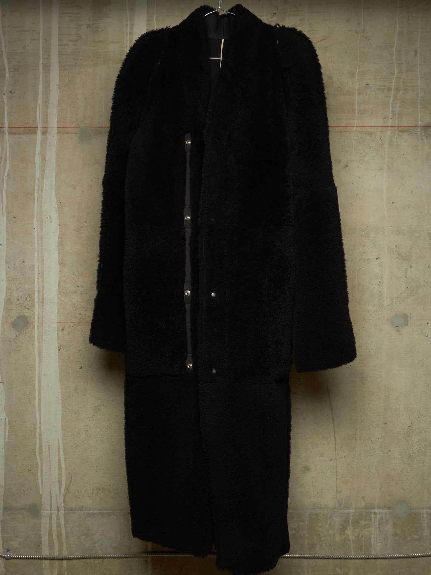 A long black teddy coat with a shawl collar and front button closure, hanging against a textured concrete wall.
