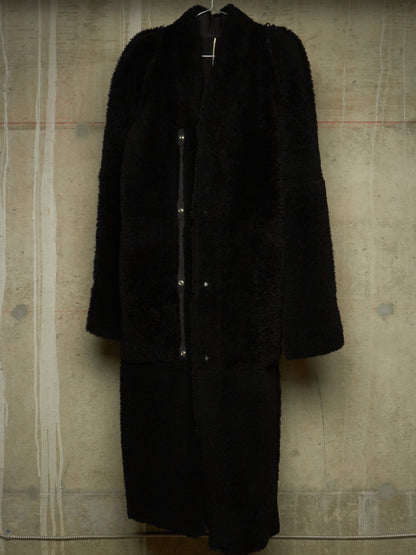 A long black teddy coat with a shawl collar and front button closure, hanging against a textured concrete wall.