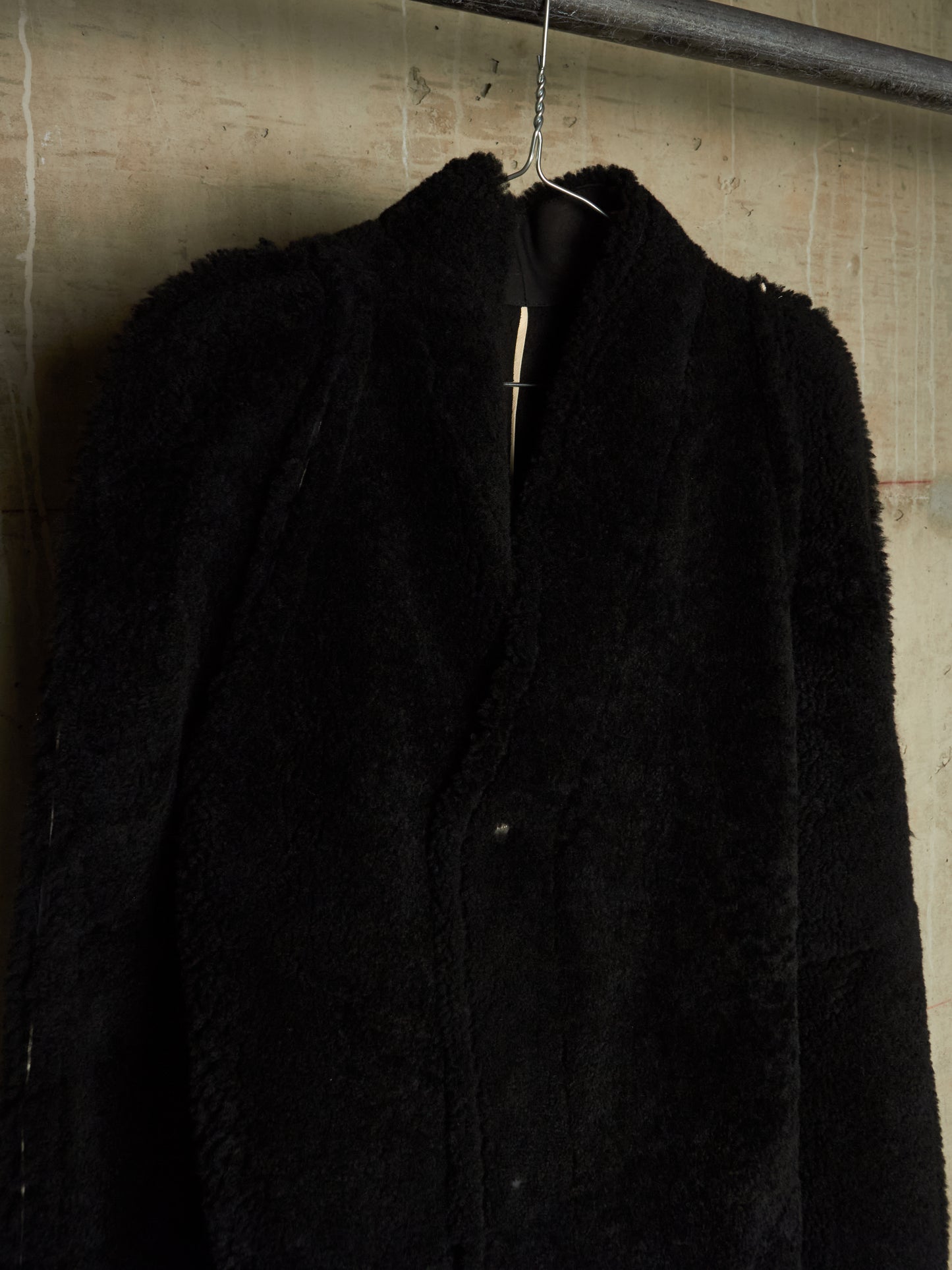 A black textured teddy coat hanging on a metal hanger against a rustic concrete wall.