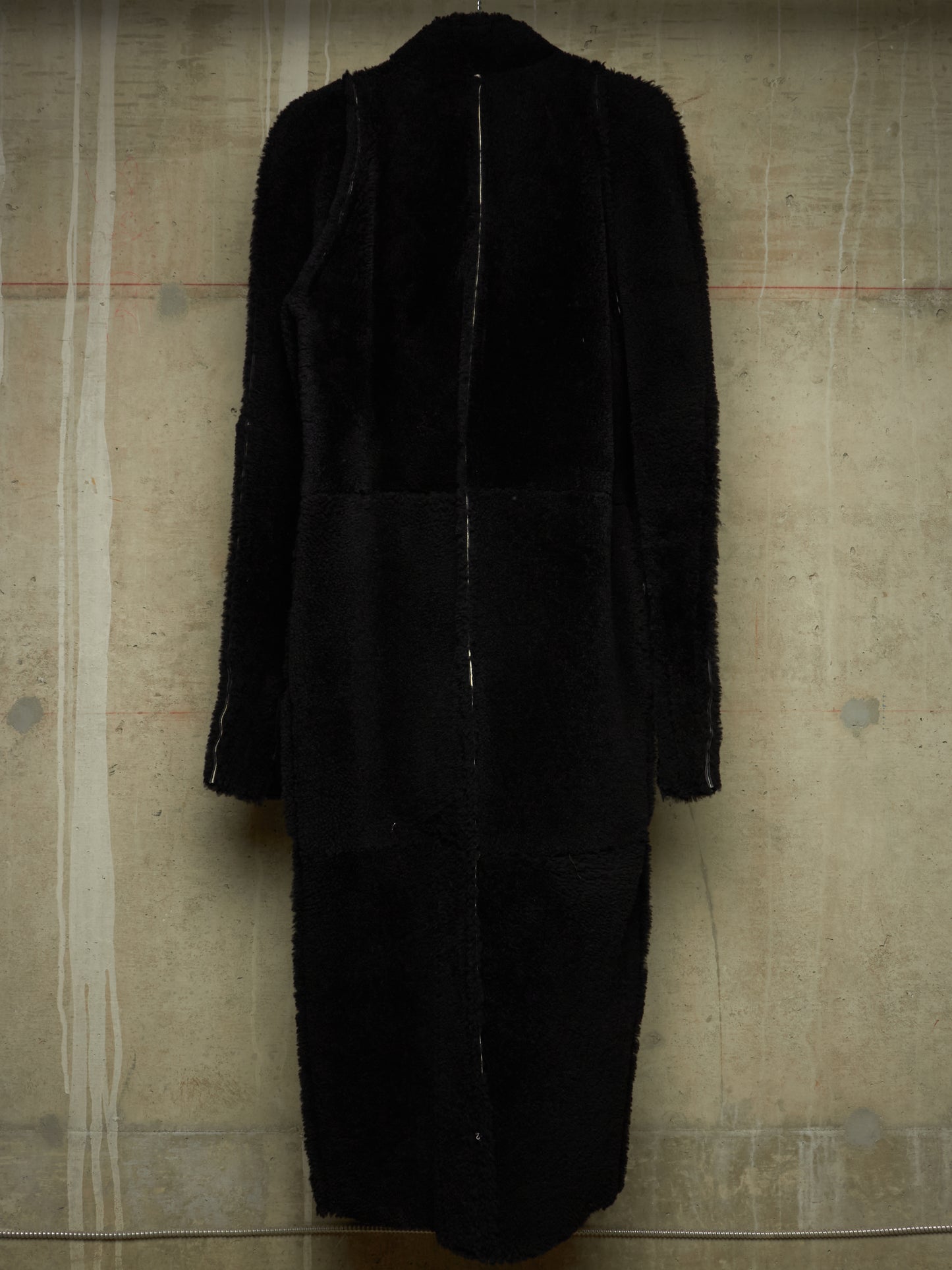 A long black coat with mixed textures, featuring a fuzzy faux-fur panel on the upper back and shoulders and smoother black fabric on the lower body, shown against a textured concrete wall.