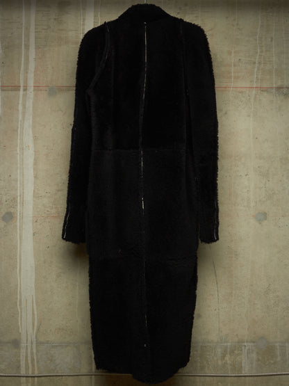 A long black coat with mixed textures, featuring a fuzzy faux-fur panel on the upper back and shoulders and smoother black fabric on the lower body, shown against a textured concrete wall.