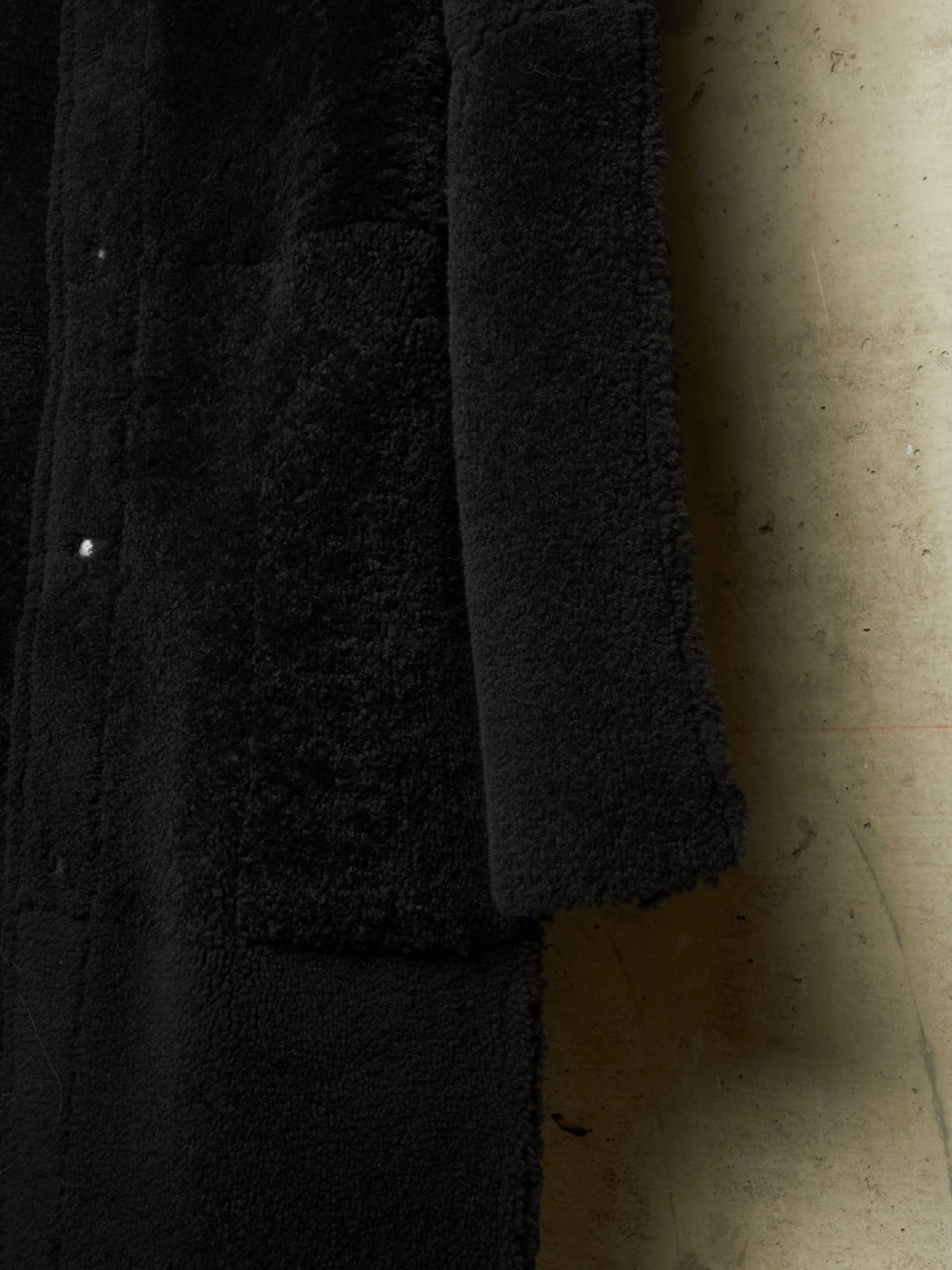 Close-up of a black textured garment, likely a coat or jacket, showing a plush sherpa or fleece lining and a smooth outer fabric, photographed against a concrete wall.