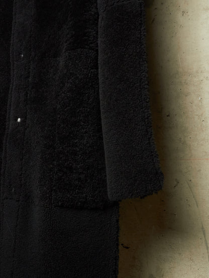 Close-up of a black textured garment, likely a coat or jacket, showing a plush sherpa or fleece lining and a smooth outer fabric, photographed against a concrete wall.