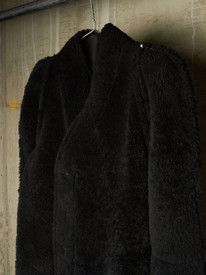 A black fuzzy teddy-fur jacket hanging on a metal hanger against a textured beige wall.