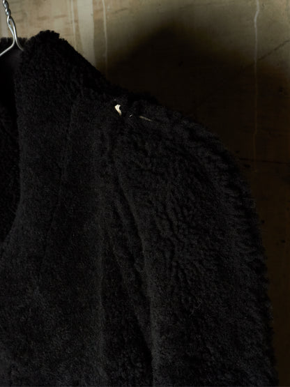 Close-up of a black textured sherpa or fleece garment hanging against a dimly lit beige wall.