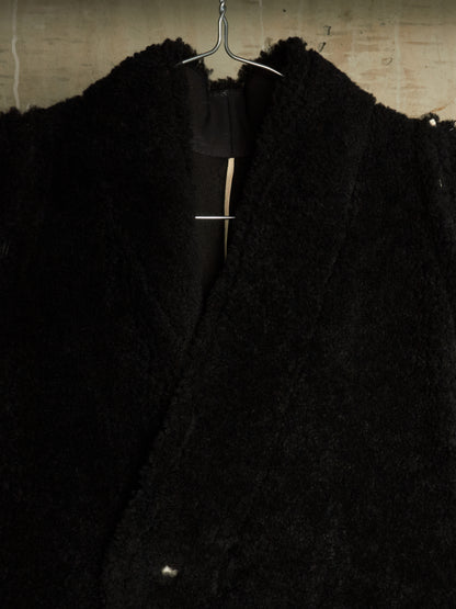 A close-up of a black textured teddy-fur or bouclé jacket hanging on a metal hanger against a light concrete wall.