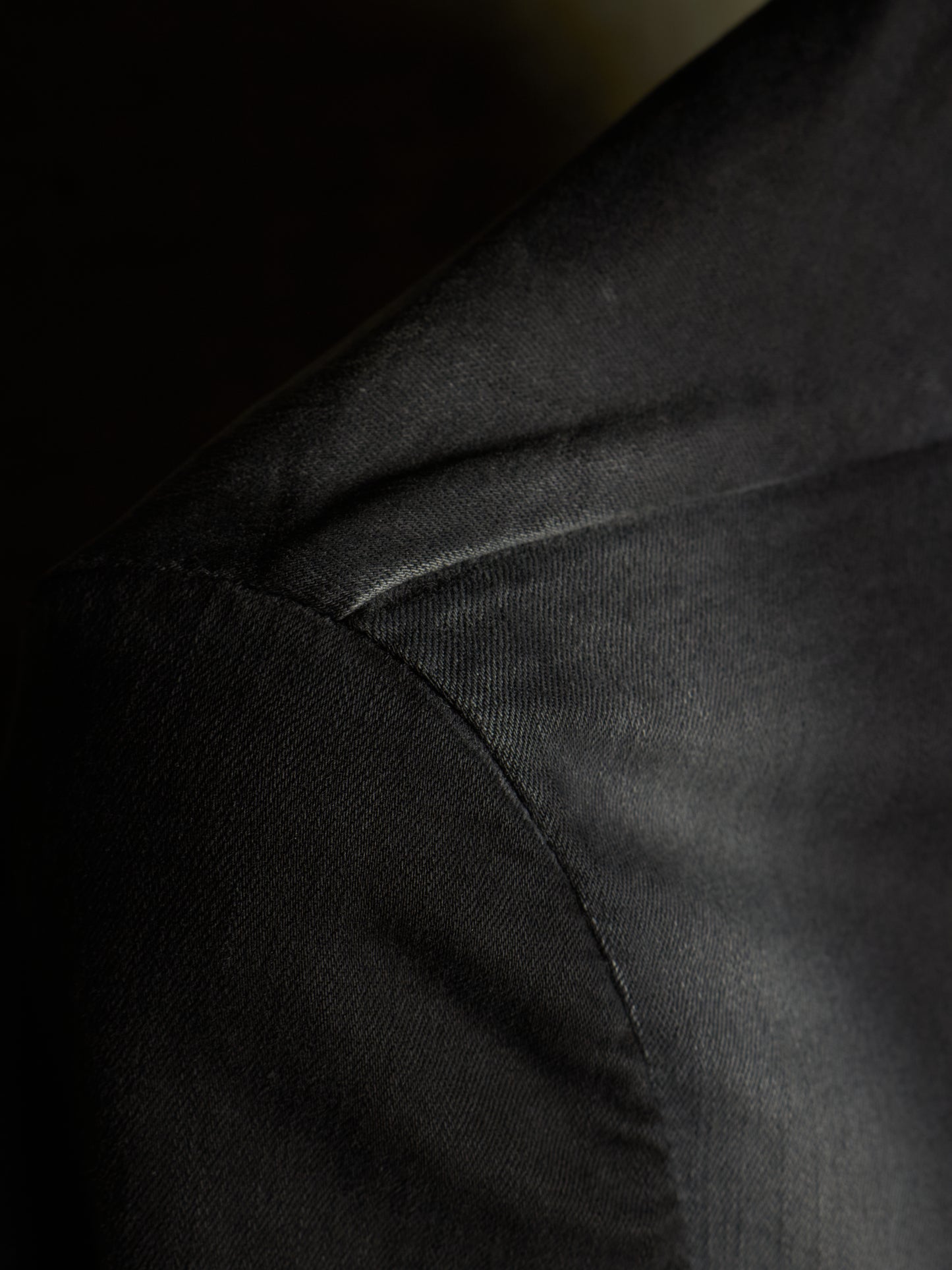 Close-up detail of a black textured fabric garment, showing the shoulder seam and fine woven texture under dramatic lighting.