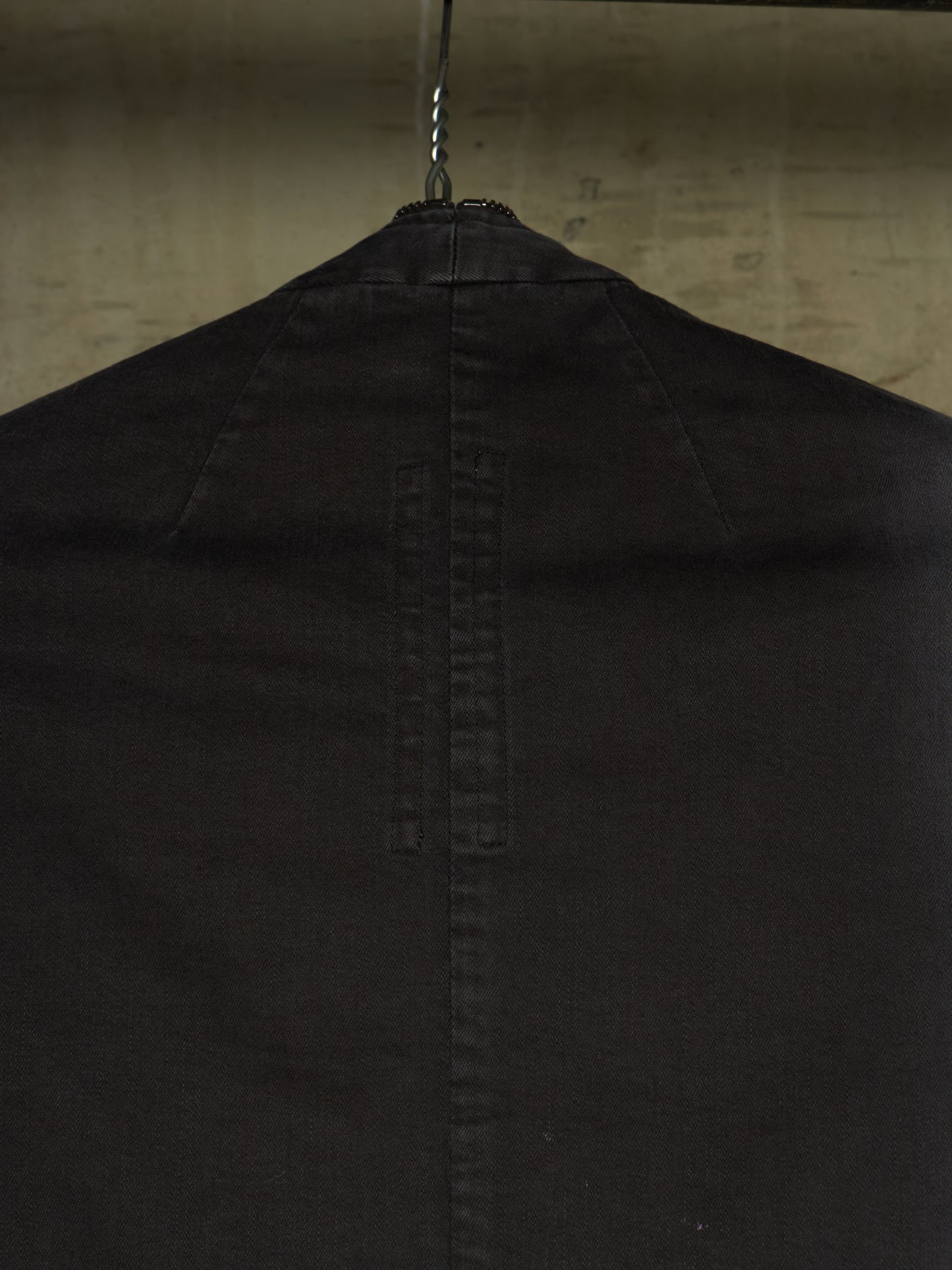 Close-up of the back of a black garment hanging on a metal hanger against a textured beige wall.