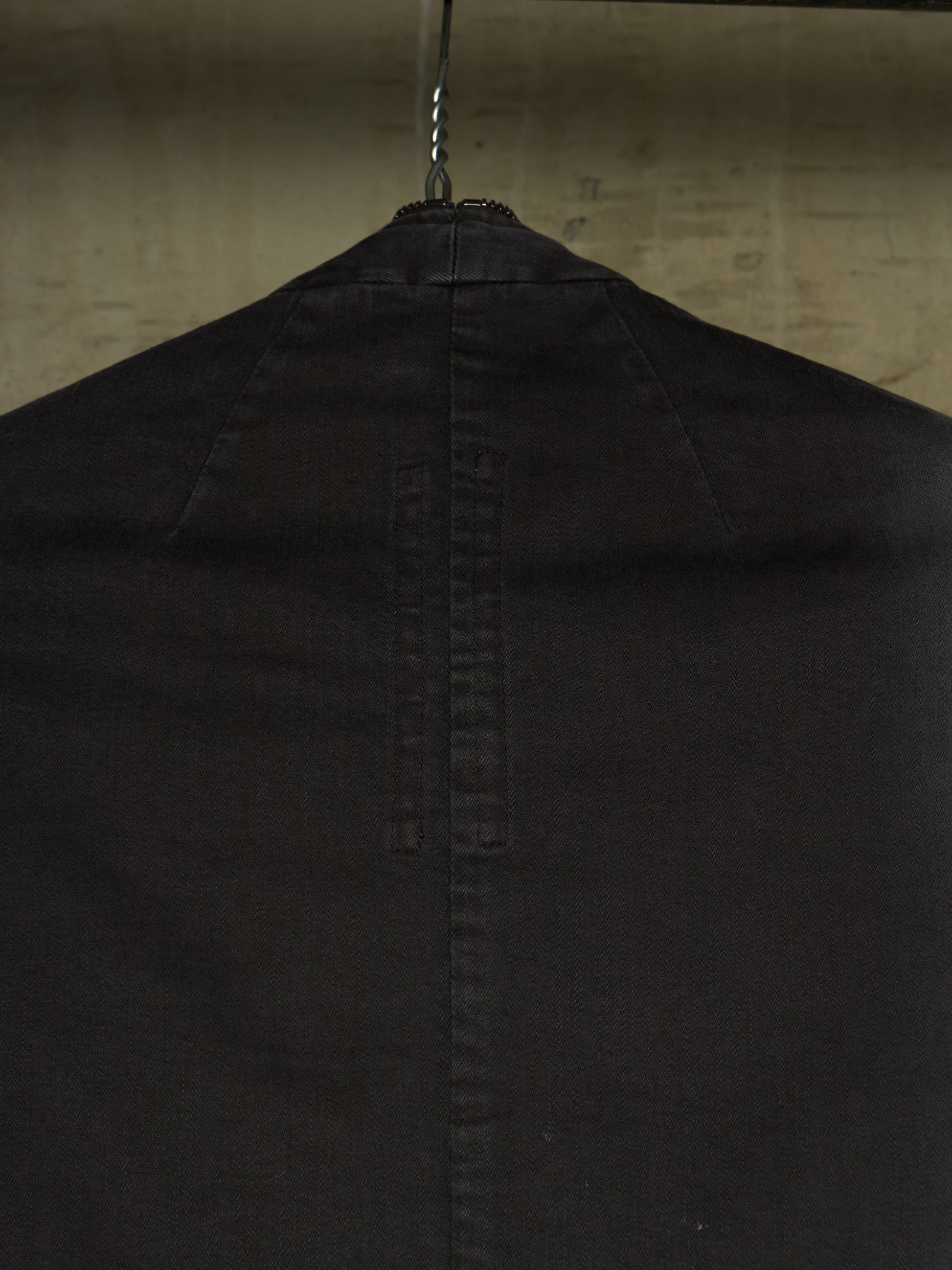 Close-up of the back of a black garment hanging on a metal hanger against a textured beige wall.